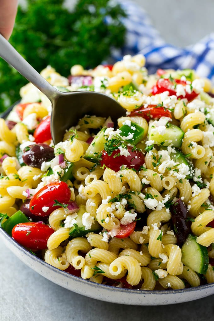 A bowl of Greek pasta salad with a serving spoon in it.